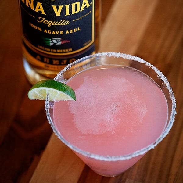 Pink margarita with a lime wedge and salted rim next to a bottle of tequila.