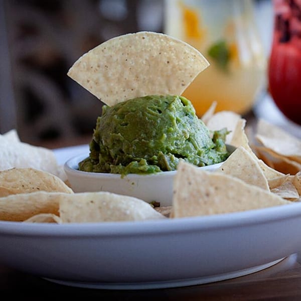 A white bowl of guacamole with a tortilla chip dipped in it, surrounded by more chips.