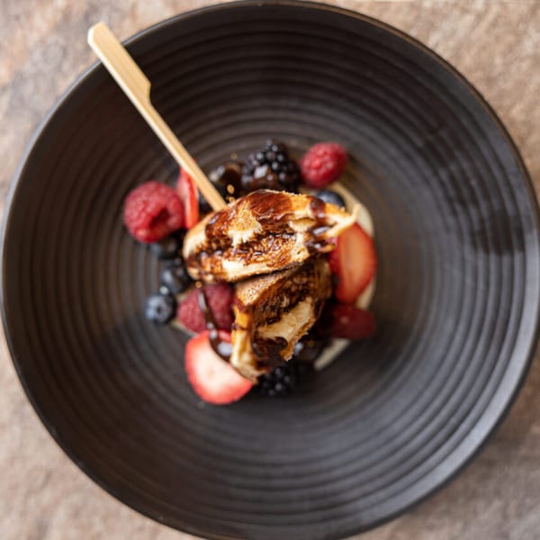 Prados' churros with chocolate drizzle, fresh strawberries, raspberries, and blueberries in a black bowl.
