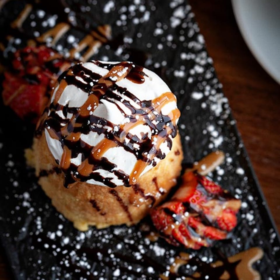 Fried ice cream dessert with whipped cream, chocolate, caramel, and strawberries on a dark plate.