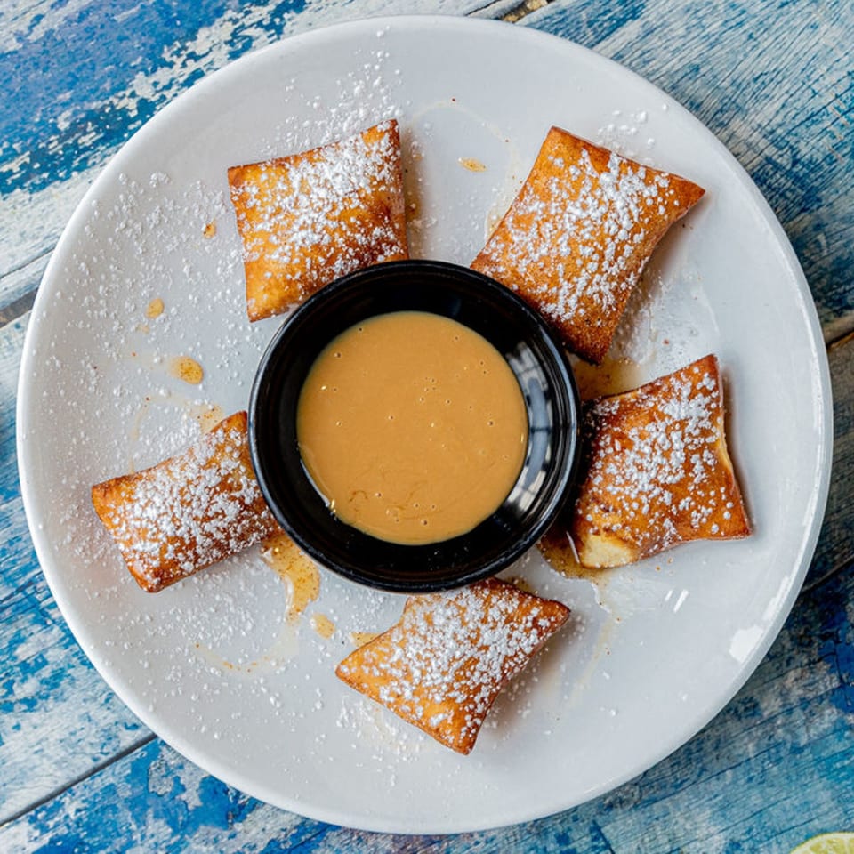Five Prados Mexican Cantina churro bites dusted with powdered sugar, surrounding a bowl of caramel sauce.