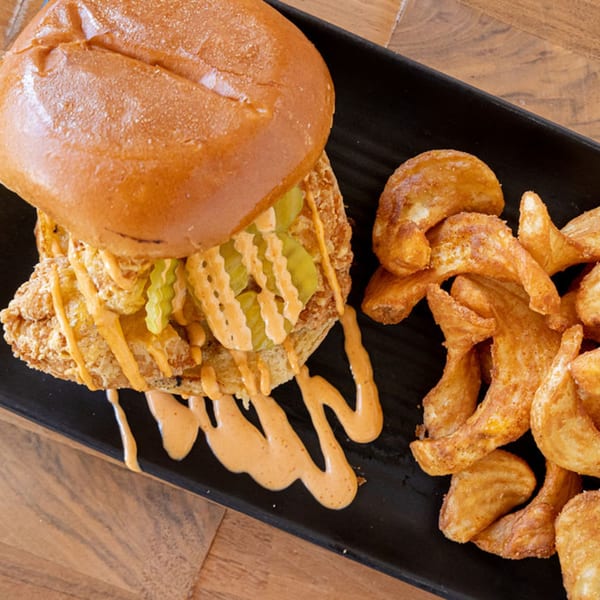 A fried chicken sandwich with pickles and sauce, served with potato wedges on a black plate.
