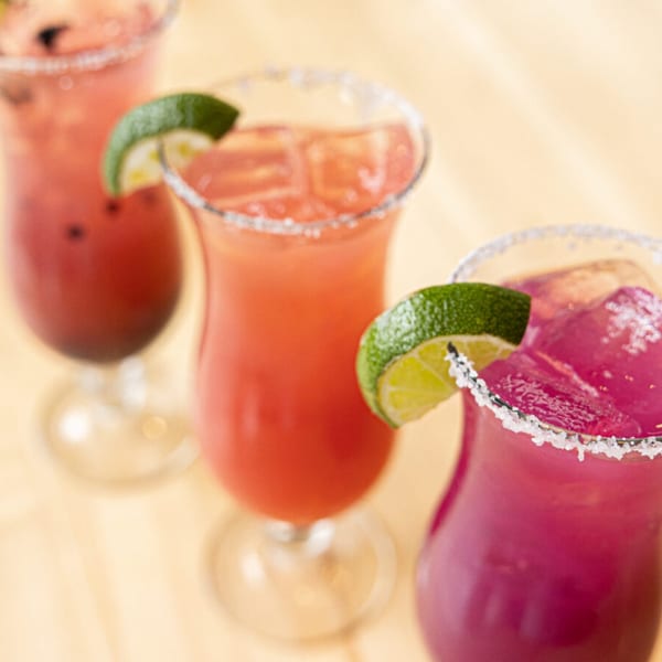 Three colorful Prados margaritas, garnished with lime wedges and salt rims, on a light wooden surface.