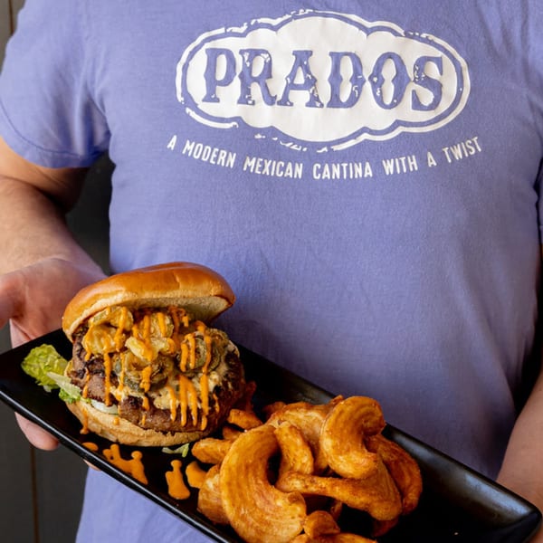 A person in a Prados Modern Mexican Cantina t-shirt holds a burger with curly fries.