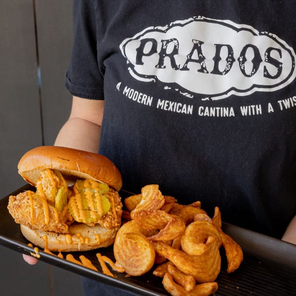 A Prados employee holds a plate with a fried chicken sandwich, pickles, sauce, and curly fries.