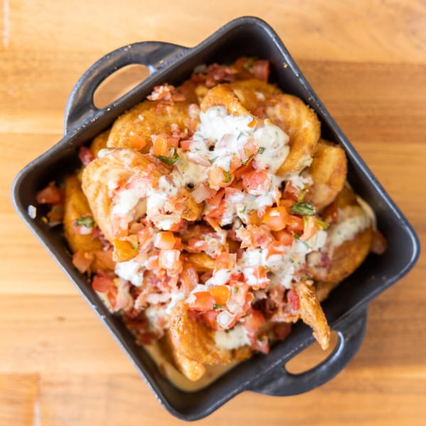 Golden fried potato wedges topped with white sauce, diced tomatoes, and herbs in a black dish.