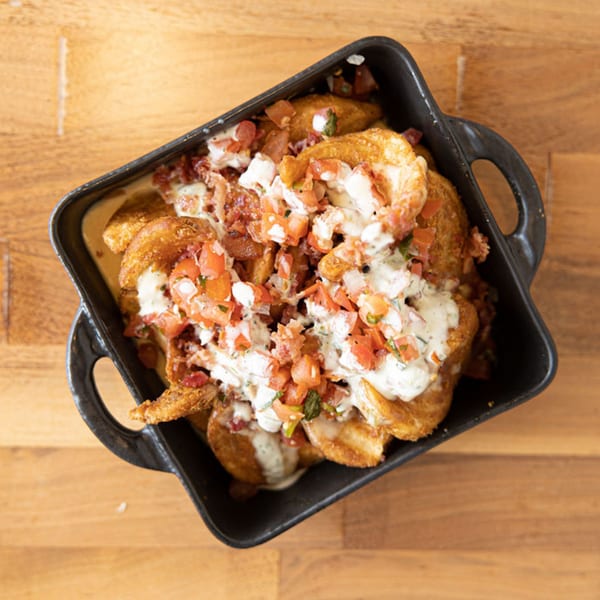 Crispy golden potato wedges topped with white sauce, diced tomatoes, and bacon bits in a black dish.