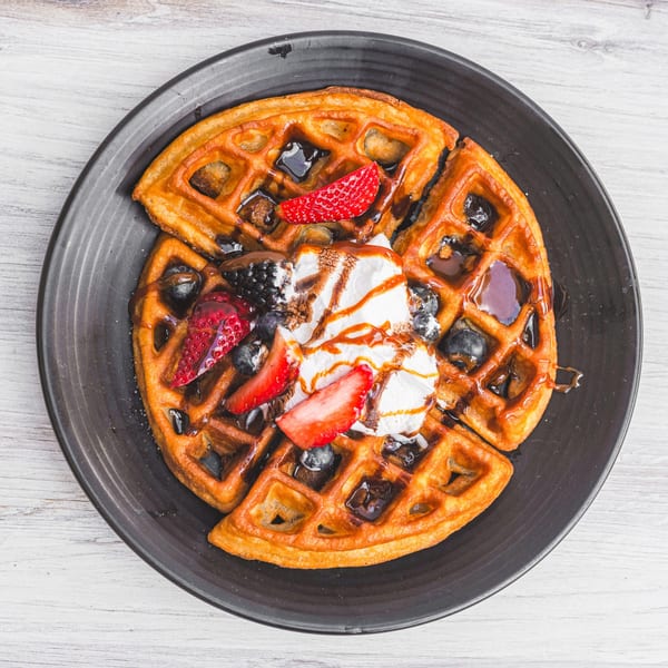 Golden waffle topped with whipped cream, strawberries, blueberries, and chocolate sauce on a black plate.