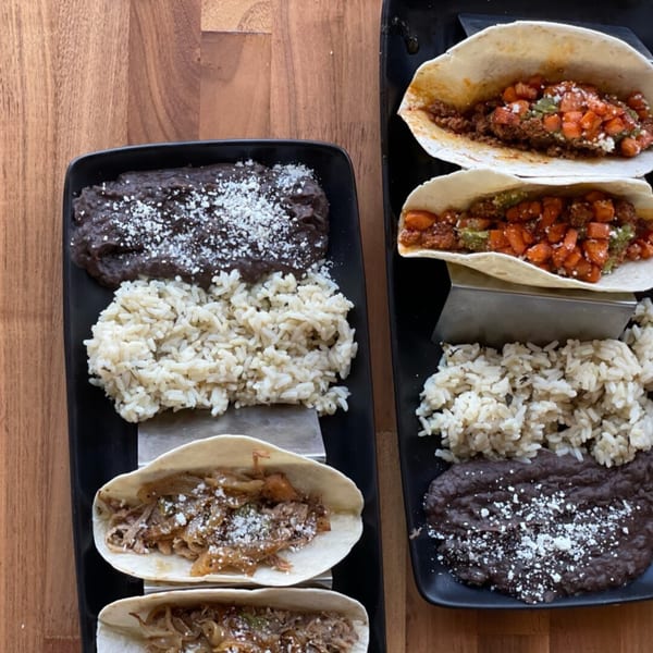 Two black plates with Prados Modern Mexican Cantina tacos, rice, and black beans on a wooden table.