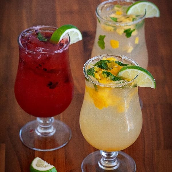 Three vibrant Prados Modern Mexican Cantina margaritas, garnished with lime, mint, and fruit, on a wooden table.