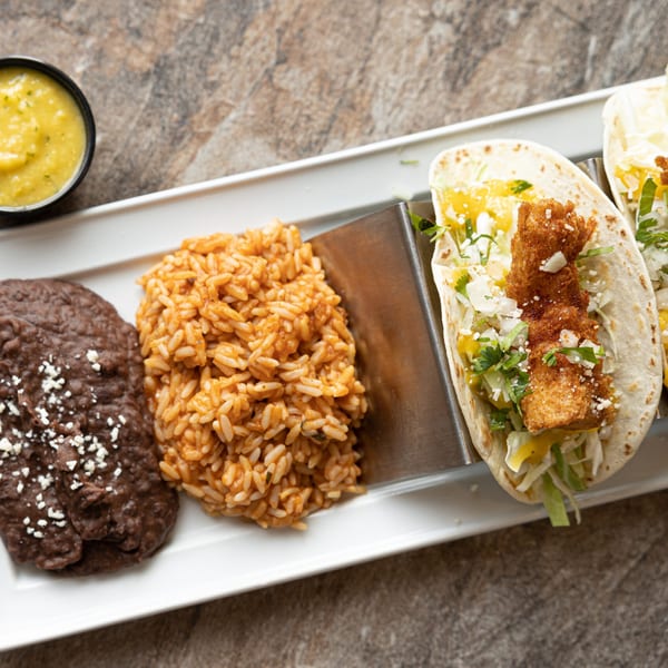 Two fish tacos with rice, beans, and a side of green salsa on a white plate.