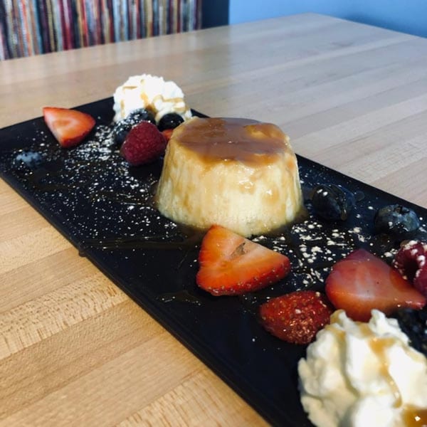 Flan dessert with caramel sauce, fresh berries, and whipped cream on a black rectangular plate.