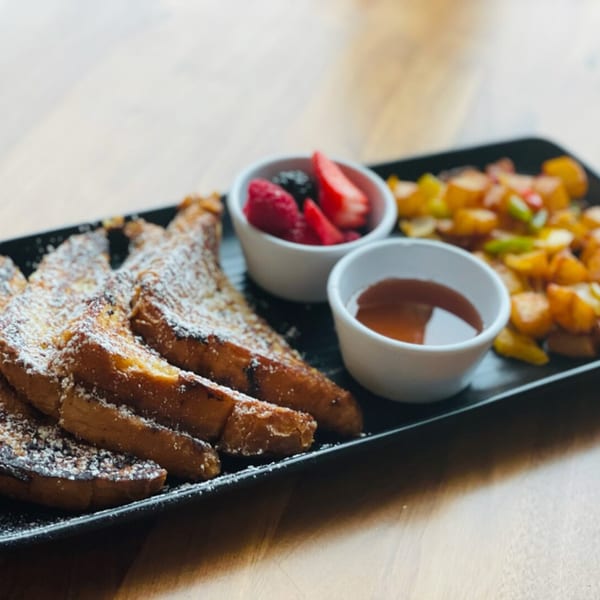 French toast with powdered sugar, berries, syrup, and breakfast potatoes on a black plate.