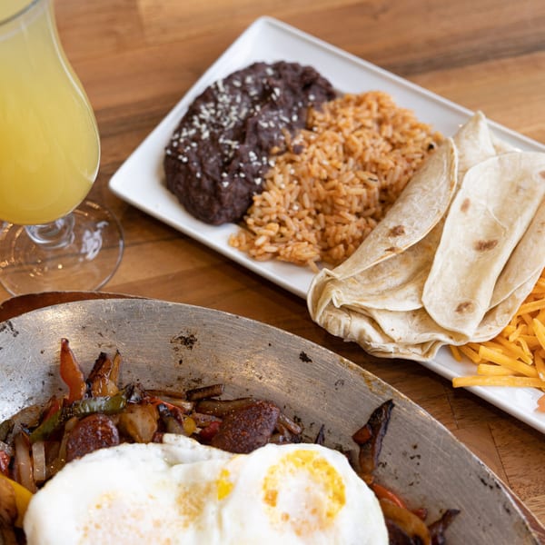 Mexican breakfast with fajitas, fried eggs, rice, beans, tortillas, shredded cheese, and a drink.