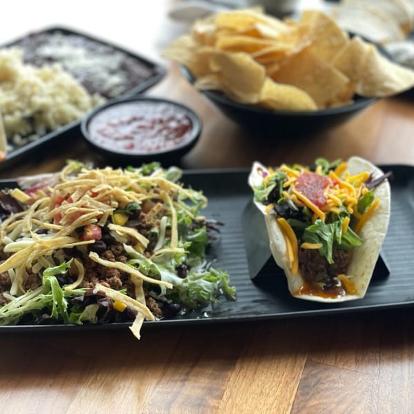 Mexican taco salad and a soft taco with chips, salsa, rice, and beans on a wooden table.